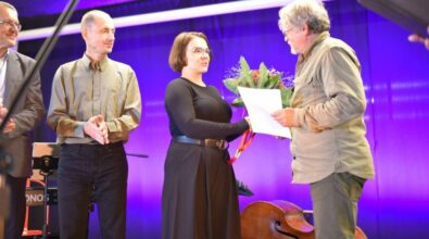 Grupka osób stoi na scenie. Kobieta ubrana na czarno wręcza laureatowi konkursu bukiet hortensji.