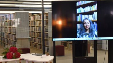 Na ekranie dużego telewizora wyświetla się kobieta. Jest to laureatka, która nie mogła osobiście zjawić się na gali. Obok stoją stoliki, na jednym z nich leży bukiet hortensji. W tle widać regały z książkami.