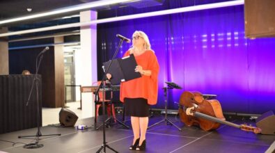 Osoba w eleganckim stroju, ubrana w czerwoną bluzkę i czarną spódnicę, przemawia na scenie. Trzyma w rękach otwartą teczkę, a obok niej stoi mikrofon. W tle widoczne są instrumenty muzyczne, w tym kontrabas, oraz fioletowe oświetlenie sceniczne.