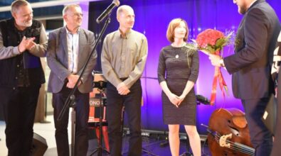 Zdjęcie przedstawia uroczyste wydarzenie, na którym jedna z osób otrzymuje kwiaty. Na scenie znajduje się grupa osób zwrócona w kierunku wręczającego kwiaty prezydenta Piotrkowa. W tle widoczne są elementy scenografii.