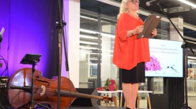 Osoba w eleganckim stroju, ubrana w czerwoną bluzkę i czarną spódnicę, przemawia na scenie. Trzyma w rękach otwartą teczkę, a obok niej stoi mikrofon. W tle widoczne są instrumenty muzyczne, w tym kontrabas, oraz fioletowe oświetlenie sceniczne.