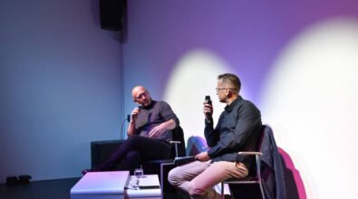 Dwóch mężczyzn siedzi na scenie przy stoliku, w rękach trzymają mikrofony i rozmawiają.