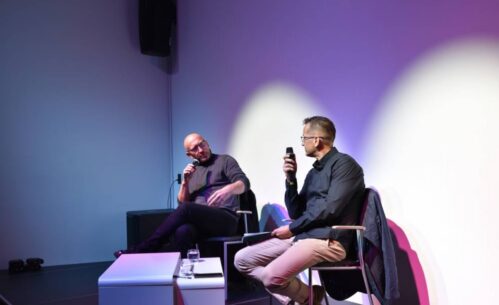 Dwóch mężczyzn siedzi na scenie przy stoliku, w rękach trzymają mikrofony i rozmawiają.