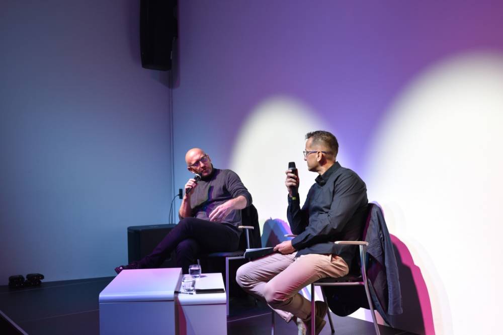 Dwóch mężczyzn siedzi na scenie przy stoliku, w rękach trzymają mikrofony i rozmawiają.