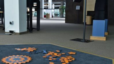 Zdjęcie przedstawia wnętrze nowoczesnego budynku. Na pierwszym planie, na dywanie, leżą rozrzucone pomarańczowe i niebieskie geometryczne kształty, częściowo ułożone w wzór przypominający mozaikę. W tle widać inne naukowe stanowiska.