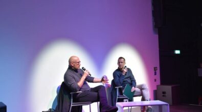 Dwóch mężczyzn siedzi na scenie przy stoliku, w rękach trzymają mikrofony i rozmawiają.