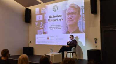 Na pierwszym planie znajduje się mężczyzna siedzący na scenie, trzymający mikrofon i przemawiający do publiczności. Za mężczyzną znajduje się duży ekran projekcyjny, na którym wyświetlane są informacje dotyczące wydarzenia. Na ekranie widoczny jest tytuł 