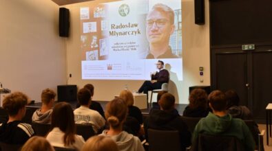 Na pierwszym planie znajduje się mężczyzna siedzący na scenie, trzymający mikrofon i przemawiający do publiczności. Za mężczyzną znajduje się duży ekran projekcyjny, na którym wyświetlane są informacje dotyczące wydarzenia. Na ekranie widoczny jest tytuł 