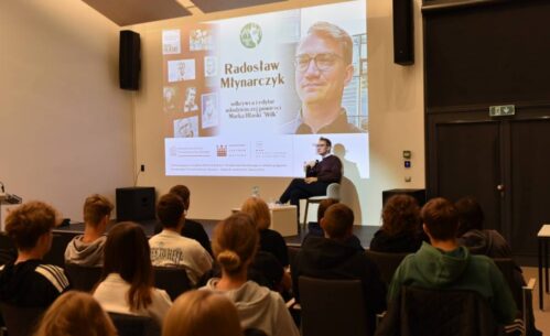 Na pierwszym planie znajduje się mężczyzna siedzący na scenie, trzymający mikrofon i przemawiający do publiczności. Za mężczyzną znajduje się duży ekran projekcyjny, na którym wyświetlane są informacje dotyczące wydarzenia. Na ekranie widoczny jest tytuł "Radosław Młynarczyk" oraz informacja, że jest on odkrywcą i redaktorem powieści młodzieżowej "Wilk" autorstwa Marka Hłaski. Ekran zawiera także kilka zdjęć Marka Hłaski oraz loga Ministerstwa Kultury i Dziedzictwa Narodowego, Narodowego Centrum Kultury oraz programu "Ojczysty - dodaj do ulubionych"