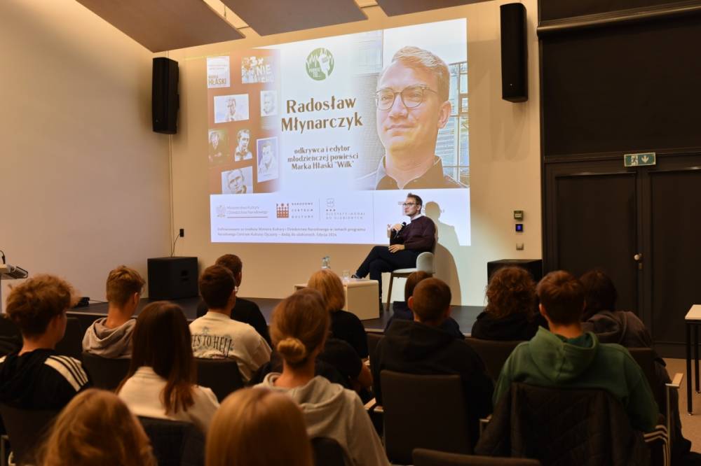 Na pierwszym planie znajduje się mężczyzna siedzący na scenie, trzymający mikrofon i przemawiający do publiczności. Za mężczyzną znajduje się duży ekran projekcyjny, na którym wyświetlane są informacje dotyczące wydarzenia. Na ekranie widoczny jest tytuł "Radosław Młynarczyk" oraz informacja, że jest on odkrywcą i redaktorem powieści młodzieżowej "Wilk" autorstwa Marka Hłaski. Ekran zawiera także kilka zdjęć Marka Hłaski oraz loga Ministerstwa Kultury i Dziedzictwa Narodowego, Narodowego Centrum Kultury oraz programu "Ojczysty - dodaj do ulubionych"