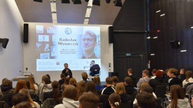 Sala pełna młodzieży, siedzi przodem do sceny. Na środku stoi kobieta przemawiająca do mikrofonu, z tyłu na scenie siedzi mężczyzna z mikrofonem. Na ekranie projekcyjnym wyświetlane są informacje dotyczące wydarzenia.