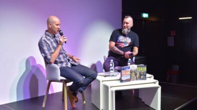 Na zdjęciu widzimy dwóch mężczyzn, siedzących na krzesłach, każdy trzyma mikrofon. Przed nimi na stole znajdują się książki.