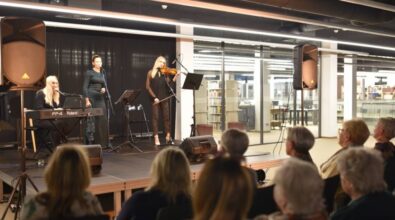 Trzy kobiety występują na scenie. Kobieta stojąca w środku śpiewa do mikrofonu, a kobiety po jej bokach grają na instrumentach. Kobieta po lewej na keyboardzie, a kobieta po prawej na skrzypcach. Widać również część publiczności.