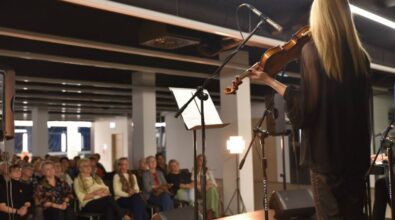 Na pierwszym planie po prawej stronie stoi kobieta grająca na skrzypcach. W tle znajduje się grupa widzów siedzących na krzesłach.