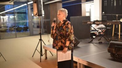 Widok na osobę prowadzącą. Kobieta w krótkich włosach ubrana w zwiewną koszulę w kwiaty przemawia do publiczności. W opuszczonej ręce trzyma kartki. W tle widać scenę przygotowaną do koncertu.