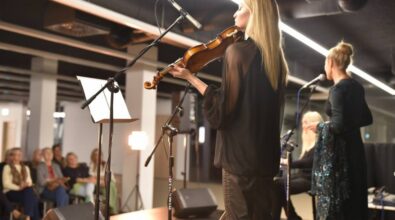 Na pierwszym planie po prawej stronie stoi kobieta grająca na skrzypcach. Przed nią stoi mikrofon, stojak z nutami, Widać także głośniki stojące na scenie. W tle znajduje się grupa widzów siedzących na krzesłach.