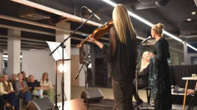 Na zdjęciu widzimy scenę podczas koncertu, na której występują muzyczki. Na pierwszym planie po lewej stronie stoi kobieta grająca na skrzypcach. Ubrana jest w czarny, zwiewny strój, a przed nią znajduje się stojak na nuty. Po prawej stronie widać drugą kobietę, śpiewającą do mikrofonu, oraz trzecią osobę grającą na fortepianie. W tle znajduje się grupa widzów siedzących na krzesłach.