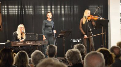 Trzy kobiety występują na scenie. Kobieta stojąca w środku śpiewa do mikrofonu, a kobiety po jej bokach grają na instrumentach. Kobieta po lewej na keyboardzie, a kobieta po prawej na skrzypcach. Widać również część publiczności. Ktoś z widowni robi zdjęcie telefonem.