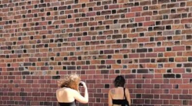 Dwie kobiety na tle murowanej ściany. Jedna kobieta stoi przodem do ściany, a druga ją fotografuje.