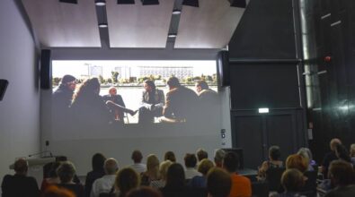 Grupa ludzi siedzi w ciemnej sali kinowej, oglądając projekcję filmu. Na ekranie widoczna jest scena rozmowy grupy osób na świeżym powietrzu, z miejską scenerią w tle, co sugeruje, że film przedstawia sceny z życia codziennego lub miejskiego. Sala jest wyposażona w nowoczesne nagłośnienie i oświetlenie, a publiczność skupiona jest na oglądanym filmie