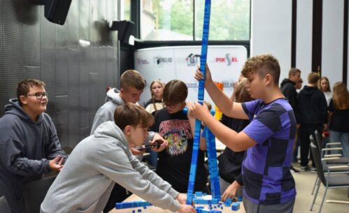 Uczestnicy budują wysoką wieżę z niebieskich klocków.