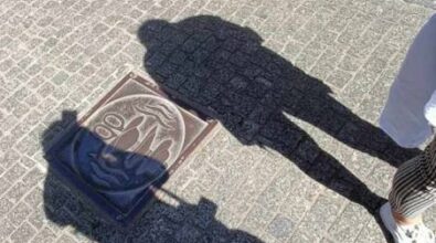 Zdjęcie przedstawia cienie dwóch osób padające na brukowaną ulicę. Na ziemi widoczna jest ozdobna płyta z symbolem nożyczek i kapeluszy. W tle widać fragment zniszczonego budynku z odpadającym tynkiem. Osoby, których cienie padają na bruk, stoją w słoneczny dzień.