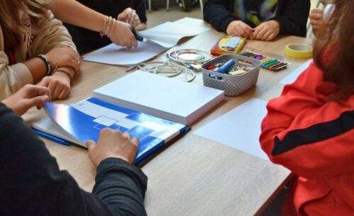 Zbliżenie na stolik uczestników, na którym leżą m.in. duży notatnik, blok, plastelina, nożyczki, taśma oraz plastikowy koszyczek z latarka i drewnianymi klockami. Dookoła widać ręce uczestników.