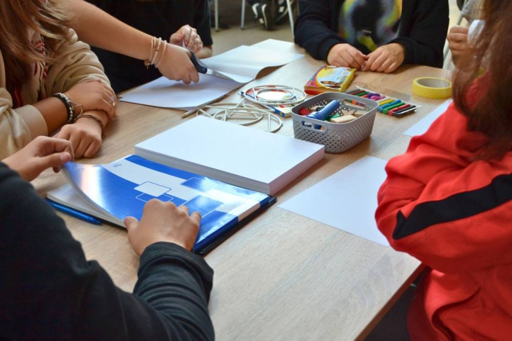Zbliżenie na stolik uczestników, na którym leżą m.in. duży notatnik, blok, plastelina, nożyczki, taśma oraz plastikowy koszyczek z latarka i drewnianymi klockami. Dookoła widać ręce uczestników.