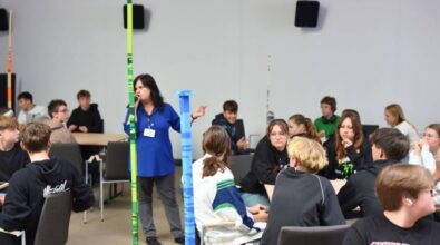 W sali pomiędzy stolikami stoją wysokie wieże w różnych kolorach. Na pierwszym planie widać wyraźnie zieloną i niebieską wieżę. W tle widać przemawiającą do mikrofonu osobę prowadzącą warsztaty.