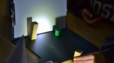 eksperyment edukacyjny z użyciem światła i cieni. Przedstawione są drewniane klocki ustawione na czarnej podstawce oraz ekran z papierem, na którym wyraźnie widać rzucane przez klocki cienie. W tle widoczna jest osoba, uczestnicząca w zajęciach.