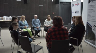 Kilka osób w różnym wieku siedzi w kręgu na krzesłach i rozmawia podczas warsztatów. Wśród uczestników są dzieci i dorośli, wszyscy zwróceni są ku sobie, co wskazuje na wspólną dyskusję. Jedna osoba siedzi tyłem do obserwatora, pozostali patrzą w jej stronę lub na siebie nawzajem. Sala jest nowoczesna, z jasnymi ścianami i ciemną okładziną na jednej z nich. Po prawej stronie ustawiony jest stojak z materiałami promocyjnymi, a w tle widać elementy wyposażenia przestrzeni edukacyjnej.