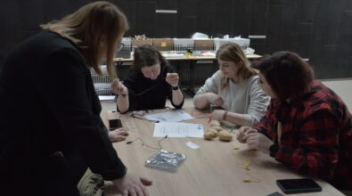 Cztery kobiety siedzą przy stole i wspólnie wykonują doświadczenie z wykorzystaniem przewodów i ziemniaków. Jedna z osób trzyma przewód i uważnie przygląda się połączeniom, pozostałe uczestniczki skupione są na montowaniu elementów układu. Na stole leżą ziemniaki, kolorowe kabelki, kartki z instrukcją oraz drobne akcesoria potrzebne do przeprowadzenia eksperymentu. Obok stoi kolejna osoba, która obserwuje przebieg pracy i wspiera uczestniczki. W tle widoczny jest blat z dodatkowymi materiałami i pomocami warsztatowymi.