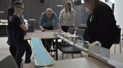 Grupa osób w różnym wieku stoi wokół stołu i obserwuje eksperyment z długą konstrukcją z papieru i taśmy, rozłożoną pomiędzy dwoma stołami. Na środku jeden z uczestników pochyla się i wprawia w ruch niewielki element, a pozostali z zaciekawieniem śledzą przebieg doświadczenia. Obok stoi chłopiec z założonymi rękami, który uważnie przygląda się całej sytuacji. Konstrukcja przypomina tor lub kanał, po którym porusza się element doświadczenia.