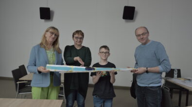 Cztery osoby – trzy dorosłe i chłopiec – stoją obok siebie i trzymają długą, papierową konstrukcję przypominającą tor. Na środku konstrukcji znajduje się mały samochodzik. Jedna z kobiet wskazuje na pojazd, pozostali uśmiechają się i patrzą w jego stronę. Wszyscy stoją w jasnej sali z prostym wyposażeniem, a w tle widoczne są krzesła, stoły oraz głośniki zamontowane na ścianie.