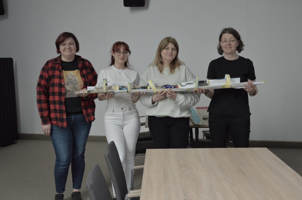 Cztery kobiety stoją obok siebie i trzymają długą, papierową konstrukcję przypominającą tor, wykonaną z kartonu, taśmy i różnych materiałów. Na środku toru znajduje się mały samochodzik. Uczestniczki uśmiechają się i patrzą w stronę obserwatora, prezentując efekt wspólnej pracy. W tle widoczne są stoły i krzesła w sali warsztatowej.