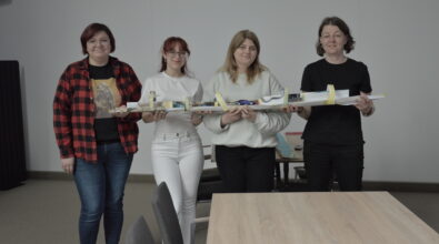 Cztery kobiety stoją obok siebie i trzymają długą, papierową konstrukcję przypominającą tor, wykonaną z kartonu, taśmy i różnych materiałów. Na środku toru znajduje się mały samochodzik. Uczestniczki uśmiechają się i patrzą w stronę obserwatora, prezentując efekt wspólnej pracy. W tle widoczne są stoły i krzesła w sali warsztatowej.