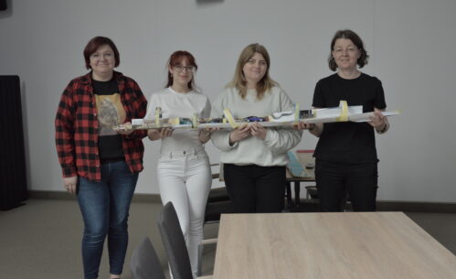 Cztery kobiety stoją obok siebie i trzymają długą, papierową konstrukcję przypominającą tor, wykonaną z kartonu, taśmy i różnych materiałów. Na środku toru znajduje się mały samochodzik. Uczestniczki uśmiechają się i patrzą w stronę obserwatora, prezentując efekt wspólnej pracy. W tle widoczne są stoły i krzesła w sali warsztatowej.