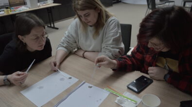 Trzy kobiety siedzą przy stole i wykonują doświadczenie z użyciem pipet oraz kropli cieczy. Każda z nich skupia się na nanoszeniu płynu na kartkę papieru, na której widać rozlane krople. Jedna z osób trzyma pipetę nad monetą lub niewielkim przedmiotem, obserwując zachowanie cieczy. Na stole leżą kartki, ołówek, chusteczka oraz telefon. Uczestniczki są pochylone nad zadaniem i uważnie obserwują efekty eksperymentu.