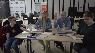 Cztery osoby – w tym chłopiec i trzy dorosłe osoby – siedzą przy stole i wykonują prace manualne z użyciem kolorowego papieru, nożyczek i innych materiałów plastycznych. Na stole znajdują się także butelki z wodą, telefon oraz różne przybory. Jedna z osób podnosi rękę, jakby zgłaszała się do odpowiedzi lub dzieliła się pomysłem, podczas gdy pozostali koncentrują się na wycinaniu i przygotowywaniu elementów. W tle widoczne są roll-upy promujące działalność instytucji oraz przestrzeń sali z ustawionymi krzesłami i stołami.