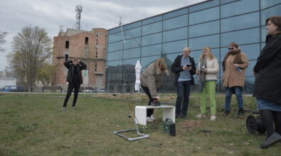Grupa osób stoi na trawie przed nowoczesnym budynkiem z przeszkloną elewacją i obserwuje doświadczenie. Jedna z osób pochyla się nad niewielkim urządzeniem ustawionym na niskim stoliku i uruchamia je, a pozostali przyglądają się z zainteresowaniem. Wokół widoczne są przewody, sprzęt oraz elementy konstrukcyjne używane do eksperymentu. Jedna osoba w tle patrzy w górę, śledząc coś unoszącego się w powietrzu. Niebo jest zachmurzone, a całość wskazuje na plenerowe zajęcia eksperymentalne.