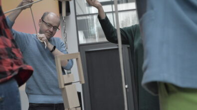 Mężczyzna trzyma sznur i reguluje ustawienie drewnianych elementów zawieszonych na konstrukcji, które tworzą układ do doświadczenia. Obok inna osoba unosi rękę, podtrzymując fragment instalacji. Drewniane klocki są zawieszone na linie i połączone z metalowym elementem, co wskazuje na eksperyment związany z ruchem lub równowagą. Wszyscy uczestnicy skupieni są na precyzyjnym ustawieniu konstrukcji.