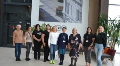 Na zdjęciu widzimy grupę bibliotekarek ustawioną w szeregu w jasnym, przestronnym wnętrzu. W tle znajduje dekoracja ścienna przedstawiająca fragment budynku. Obok widoczne są rośliny doniczkowe.