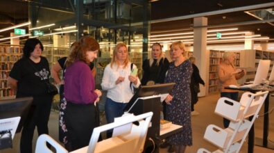 Na zdjęciu widać grupę osób zgromadzonych wokół stanowiska z komputerem. W tle znajdują się regały z książkami oraz nowoczesne, przestronne wnętrze biblioteki.