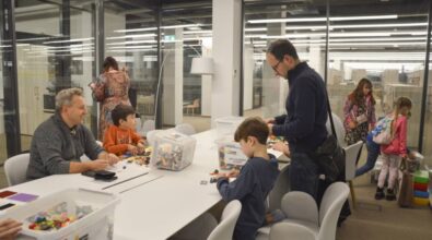 Na zdjęciu przedstawiona jest sala z klockami LEGO. Przy dużym białym stole siedzą dzieci wraz z dorosłymi, zajęci budowaniem z klocków, które znajdują się w przezroczystych pojemnikach rozmieszczonych na stole. Po prawej stronie widoczny jest mężczyzna pomagający dziecku, a w tle inne dzieci przeglądają dostępne materiały. W pomieszczeniu panuje przyjazna atmosfera, a przez szklane ściany można dostrzec kolejne przestrzenie wypełnione światłem. Całość oddaje klimat twórczej zabawy i współpracy międzypokoleniowej.