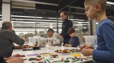 Noc Bibliotek 2024. Duży stół w przeszklonej sali, na którym rozsypane są klocki lego. Na stole widać przechylone plastikowe pudełka, z których wysypują się klocki. Przy stole siedzą dorosłe osoby oraz dzieci i budują różne konstrukcje. Jeden mężczyzna oraz chłopiec stoją przy stole.