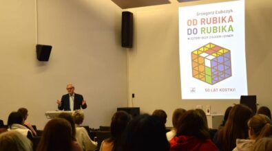 Grzegorz Łubczyk, starszy mężczyzna w garniturze i okularach przemawia do publiczności. Na ekranie widać zdjęcie jego książki „Od Rubika do Rubika”, przedstawiającą kolorową kostkę Rubika i świętującą 50 lat.