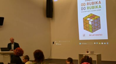 Uśmiechnięty autor przemawia do publiczności. Na ekranie widać zdjęcie książki „Od Rubika do Rubika”, przedstawiającą kolorową kostkę Rubika i świętującą 50 lat.