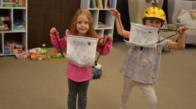 Na zdjęciu widać dwoje dzieci stojących w pokoju, każde z nich trzyma plecak ściągany sznurkiem z nadrukowanym wzorem zamku i chmur. W tle znajdują się półki wypełnione książkami i grami planszowymi, a także zabawki na podłodze, w tym zestaw kolejki i kolorowa zabawka z piłką.