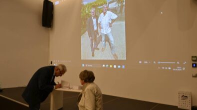 Autor podpisuję książkę pochylając się nad stolikiem stojącym na scenie. W tle widać zdjęcie z prezentacji. Obok stoi starsza kobieta czekająca na książę.