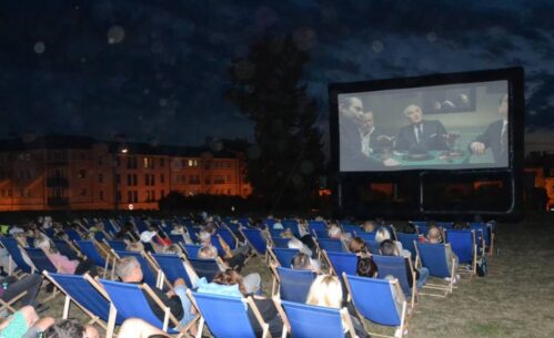 Ludzie siedzący na leżakach oglądają film na dużym telebimie. Jest wieczór, w oddali widać oświetlone budynki.